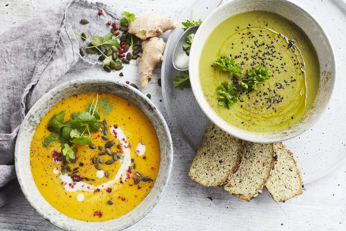 Soup in Lebanon, Ready Soups, Healthy Soups in Lebanon, Two ceramic bowls of soup on a light gray surface, one orange-yellow and one green-yellow, garnished with herbs, seeds, and cream, with slices of bread and ginger nearby.