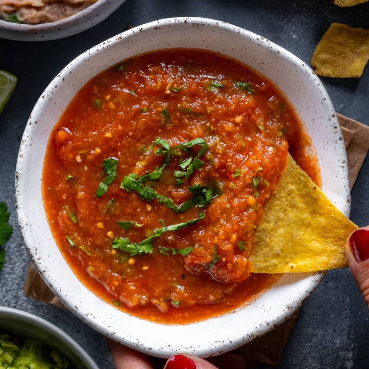 A bowl of chunky red salsa with cilantro garnish, with a hand dipping a tortilla chip into the salsa. Surrounding props include a lime wedge, guacamole, beans, and scattered tortilla chips on a dark tabletop.