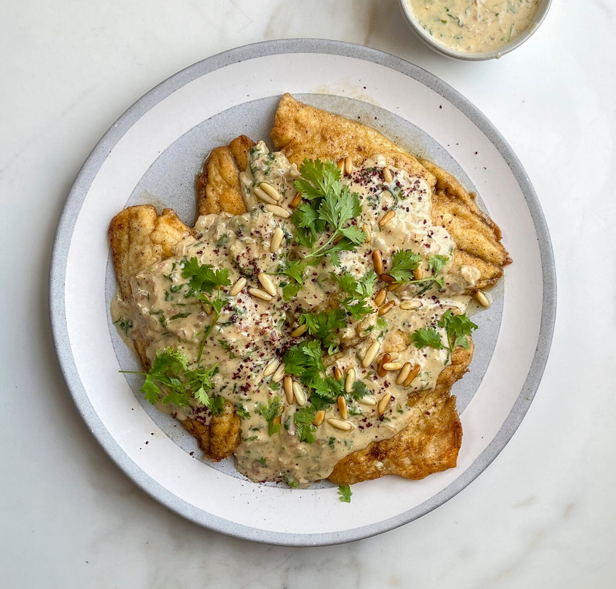 A top-down view of a plated food dish on a white marble surface, featuring golden-brown fried or baked pieces covered in a creamy beige sauce and topped with chopped green herbs, with a small side bowl of dressing.