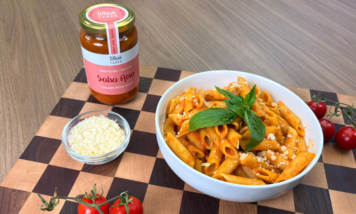 A bowl of tomato-based penne pasta garnished with fresh basil and grated cheese, shown on a wooden checkered board with a jar of Salsa Rosa pasta sauce and a small bowl of cheese.

