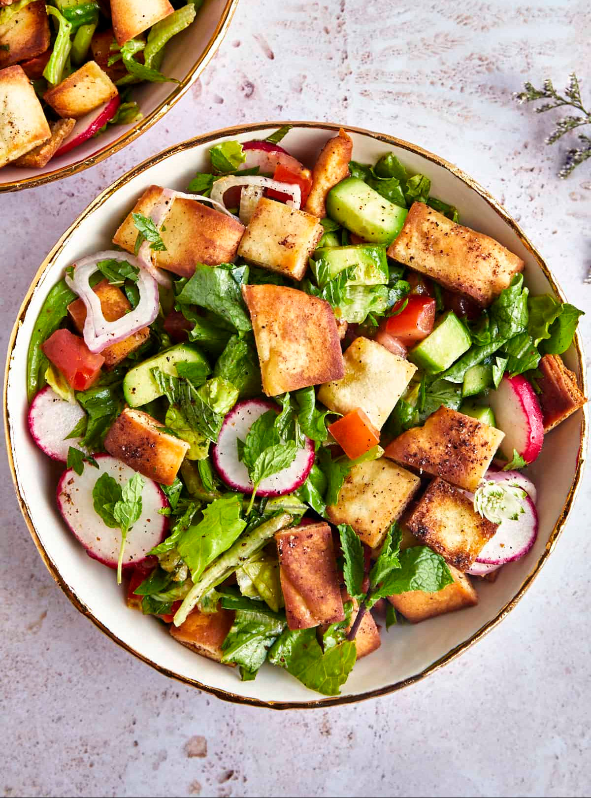 A top-down view of a fattoush in a white bowl with a gold rim, featuring golden-brown croutons, mixed greens, radishes, cucumbers, tomatoes, and dressing, with a second bowl partially visible in the upper left on a light textured surface. Lebanese Food Fattoush Dressing Healthy Whisktaker