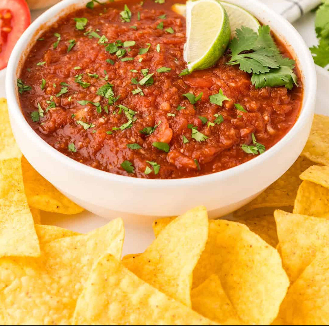A white bowl filled with red salsa garnished with chopped cilantro, a lime wedge, and parsley, surrounded by tortilla chips, with a whole onion, tomato slice, and checkered cloth in the background.