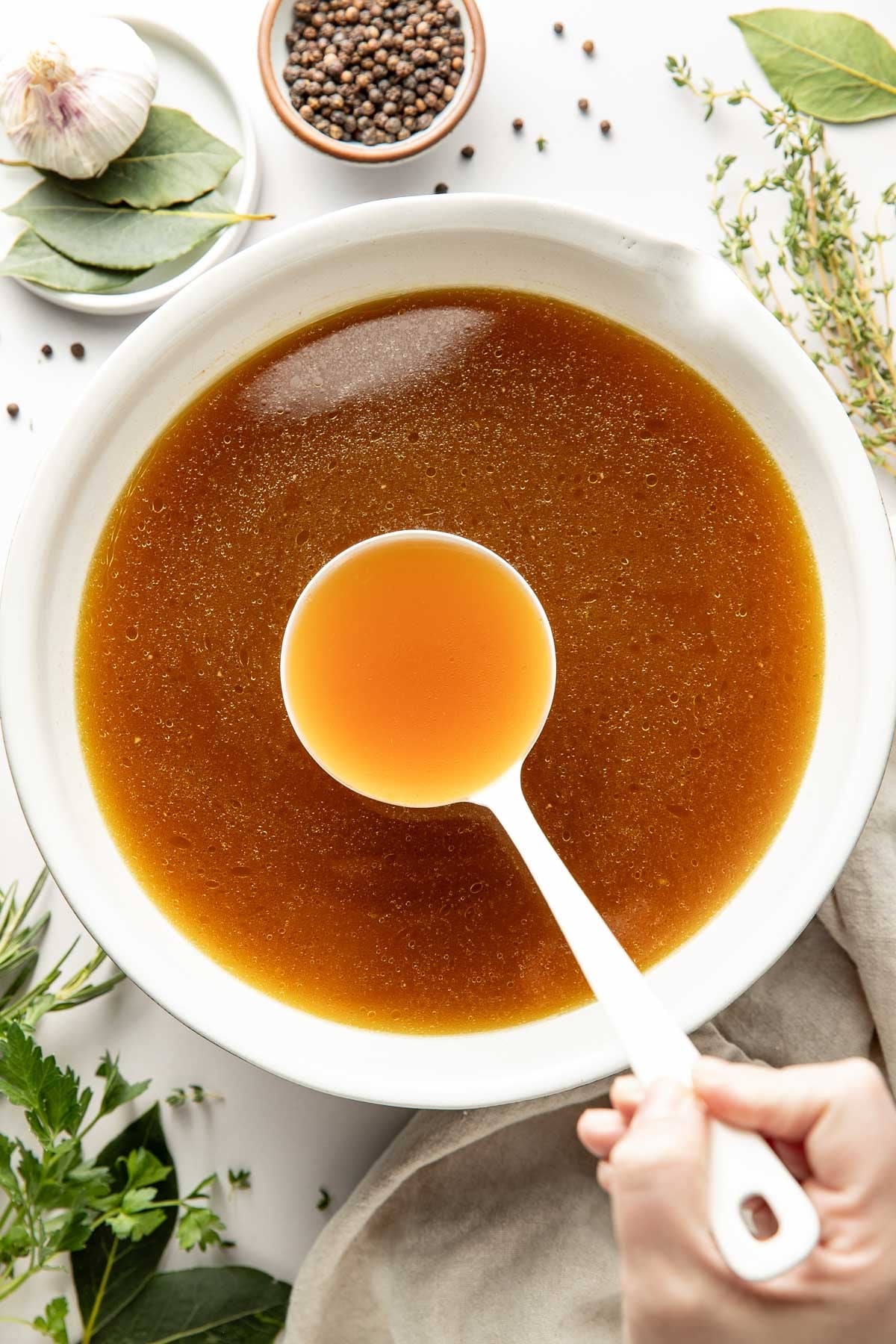 Bone Broth in Lebanon. Collagen rich. A top-down view of a white bowl filled with amber-brown broth or stock, with a hand holding a white ladle above the bowl. Fresh herbs, a garlic bulb, and a small bowl of peppercorns are arranged around the bowl on a light surface.