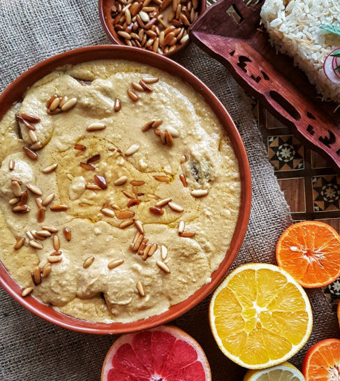 ARENBIYEH - KIBBEH ARENBIYEH A styled overhead food scene featuring a large bowl of creamy beige dip or spread topped with sunflower seeds, a tray of white rice, and sliced citrus fruits including orange, grapefruit, lime, and tangerine on a textured fabric surface.